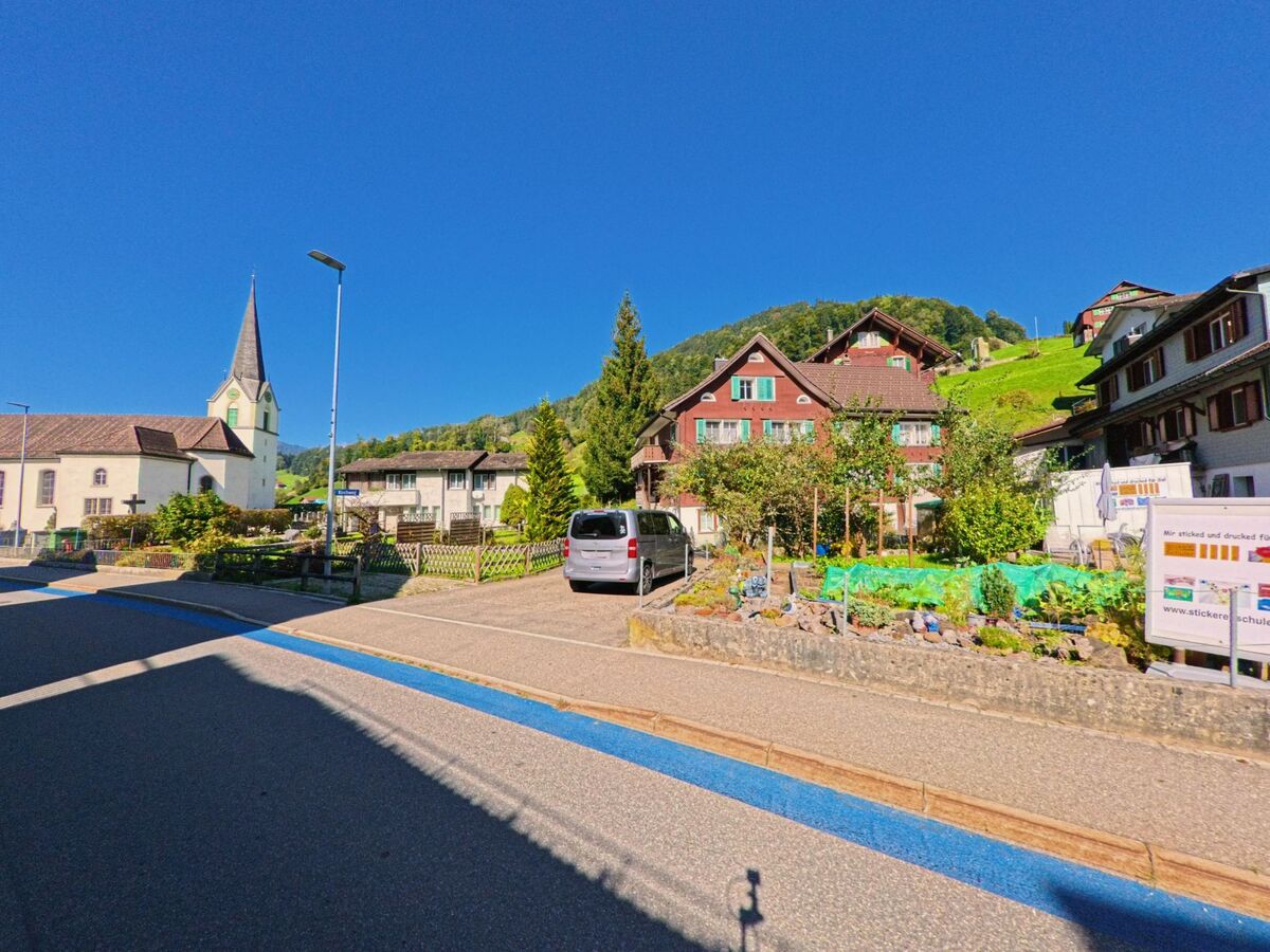 In Vorderthal (8857, Kanton Schwyz) zeigt das Bild einen kleinen Gartenbereich vor traditionellen Holzhäusern, während links die Kirche am Rand der Strasse steht.