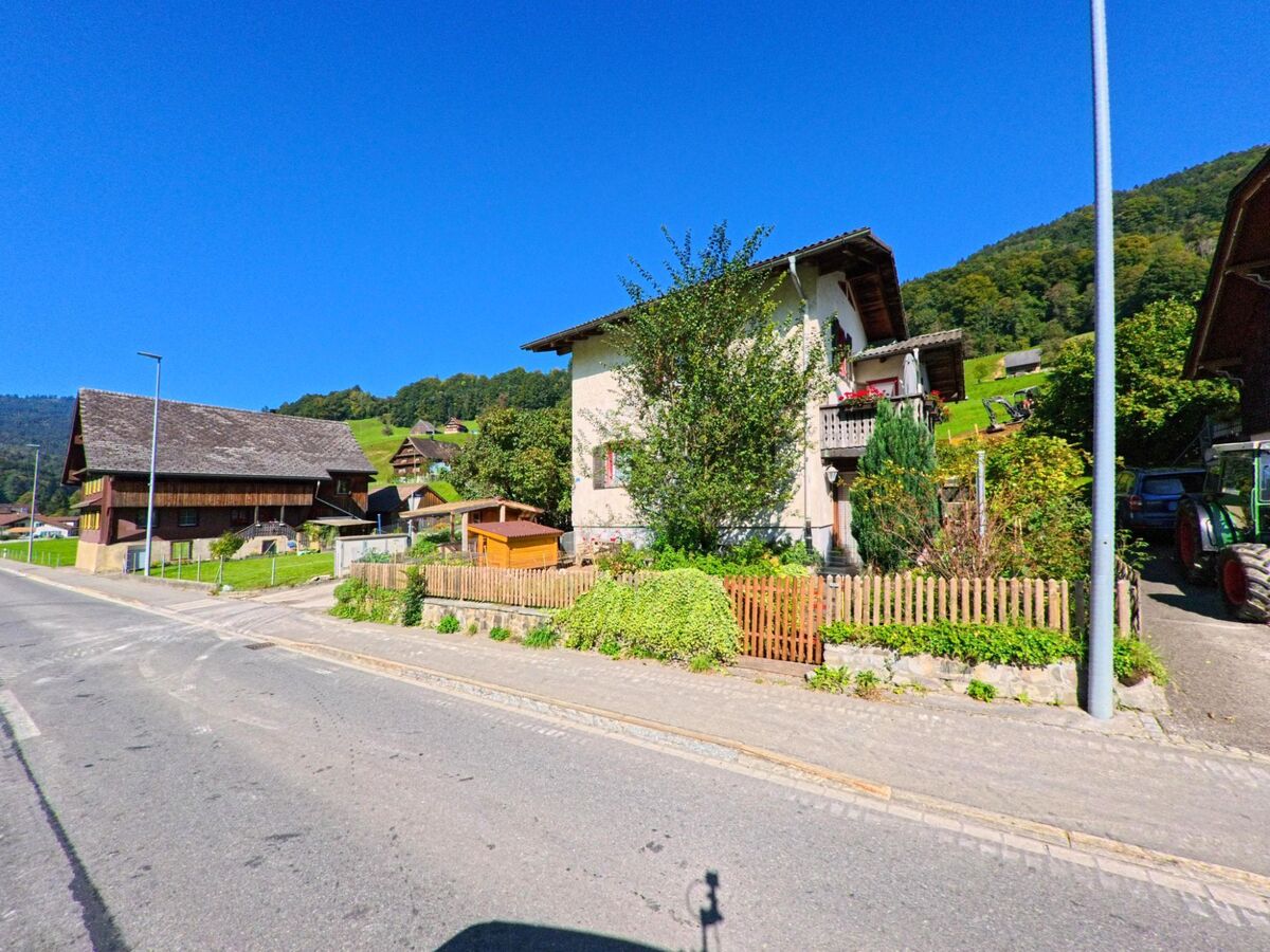 In Vorderthal (8857, Kanton Schwyz) stehen mehrere Haeuser mit Holzelementen und kleinen Gaerten entlang der Strasse, waehrend im Hintergrund der bewaldete Hang ansteigt.