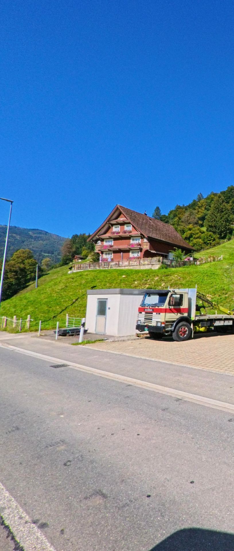 In Vorderthal (8857, Kanton Schwyz) steht ein traditionelles Holzhaus mit Blumenkasten am Hang, waehrend im Vordergrund ein alter Lastwagen neben einem kleinen Gebaeude parkiert ist.