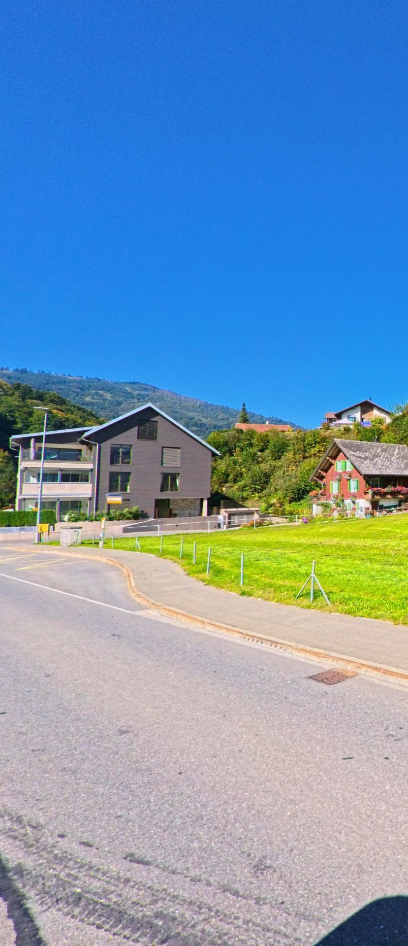 In Vorderthal (8857, Kanton Schwyz) stehen ein modernes Mehrfamilienhaus und ein traditionelles Holzhaus vor einem Hang, der mit dichter Vegetation und einzelnen Gebaeuden durchzogen ist.