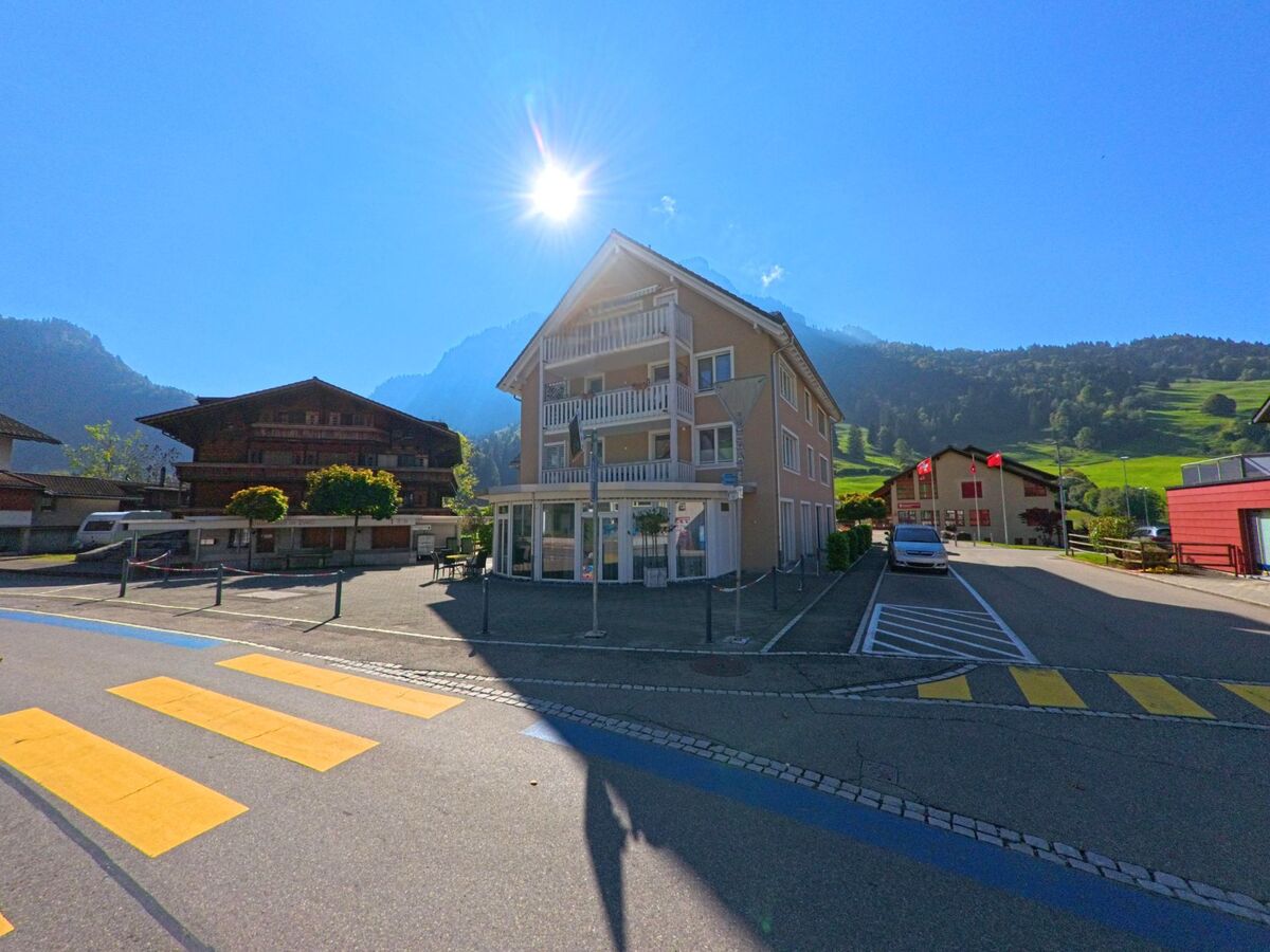 In Vorderthal (8857, Kanton Schwyz) faellt die Sonne hell auf ein modernes Wohnhaus mit Balkonen, das zwischen traditionellen Holzbauten und den ansteigenden Gruenhaengen steht.