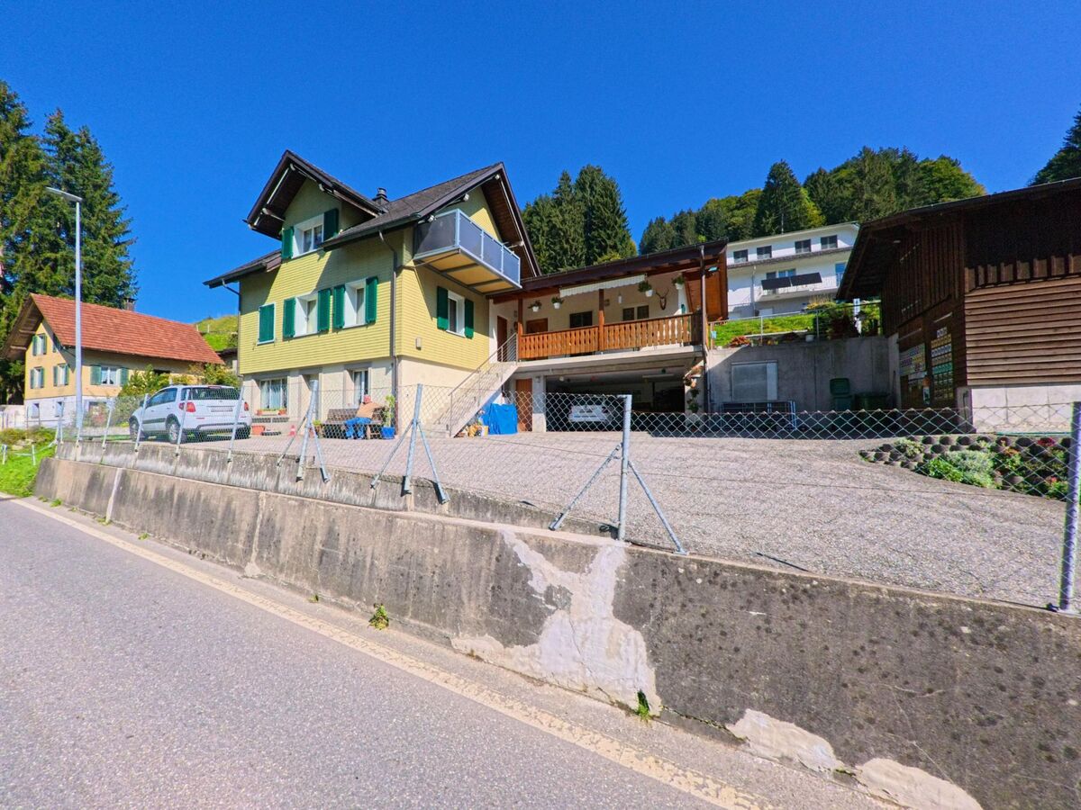 In Vorderthal (8857, Kanton Schwyz) stehen mehrere Wohnhaeuser mit Holzverkleidung und Balkonen an einem ansteigenden Hang, umgeben von Wald und einzelnen Gaerten.