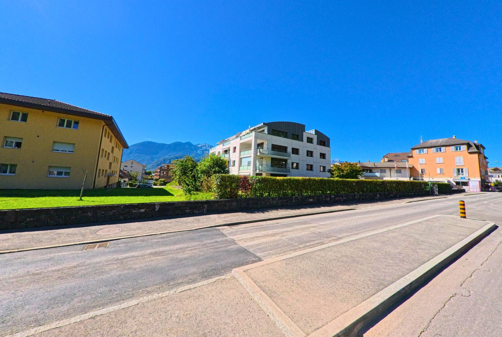 Vor modernen Wohnhaeusern in Vouvry (1896, Kanton Wallis) fuehrt eine breite Strasse entlang gepflegter Gruenflaechen mit Blick auf die Berge im Hintergrund.