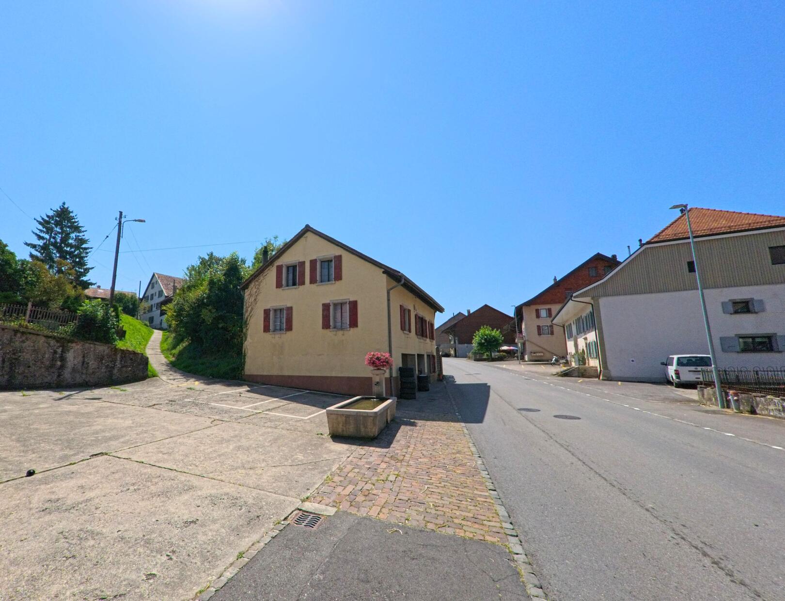 Ein kleines Haus mit roten Fensterlaeden steht an einer ansteigenden Strasse im Dorf Vuarrens (1418, Kanton Waadt).