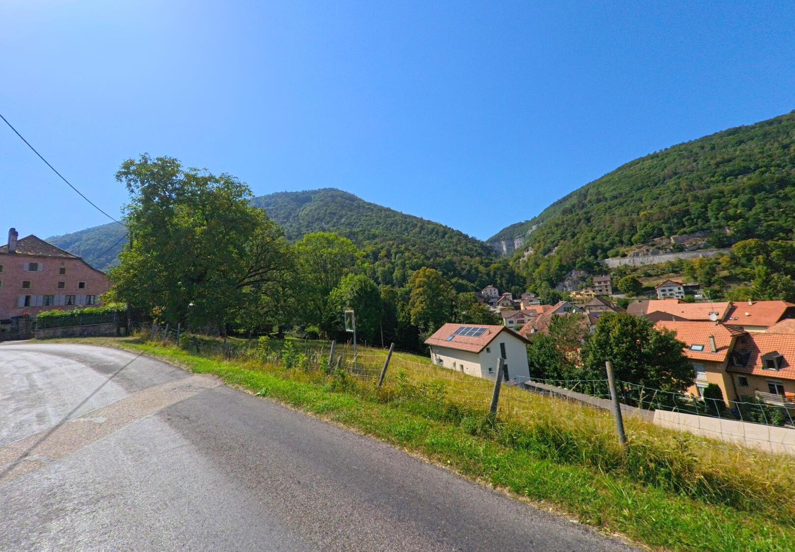 Von einer ansteigenden Strasse in Vuiteboeuf (1445, Kanton Waadt) bietet sich ein Blick auf die Haeuser des Dorfes, das eingebettet zwischen bewaldeten Huegeln liegt.