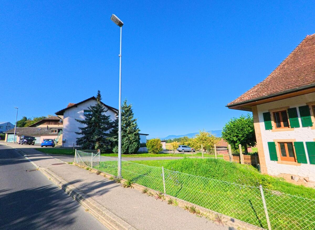 In Vully-les-Lacs (1589, Kanton Waadt) stehen Wohnhaeuser mit gruene Fensterlaeden und gepflegten Gaerten entlang einer leicht ansteigenden Strasse.