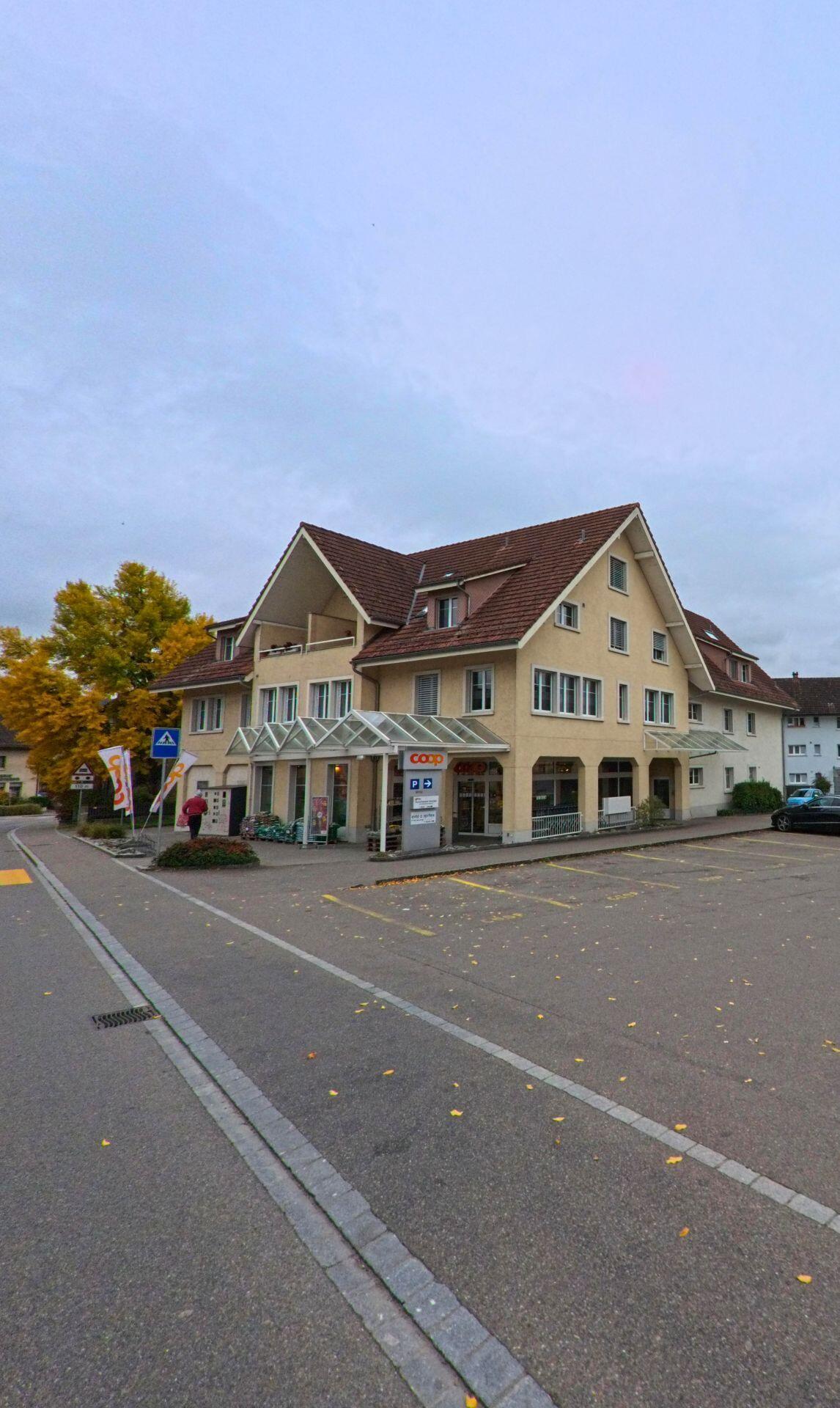 Vor einem Einkaufsgebäude mit Glasvordach und Parkplatz zeigt sich ein zentraler Versorgungsstandort in Wängi (9545, Kanton Thurgau) an einem ruhigen Herbsttag.