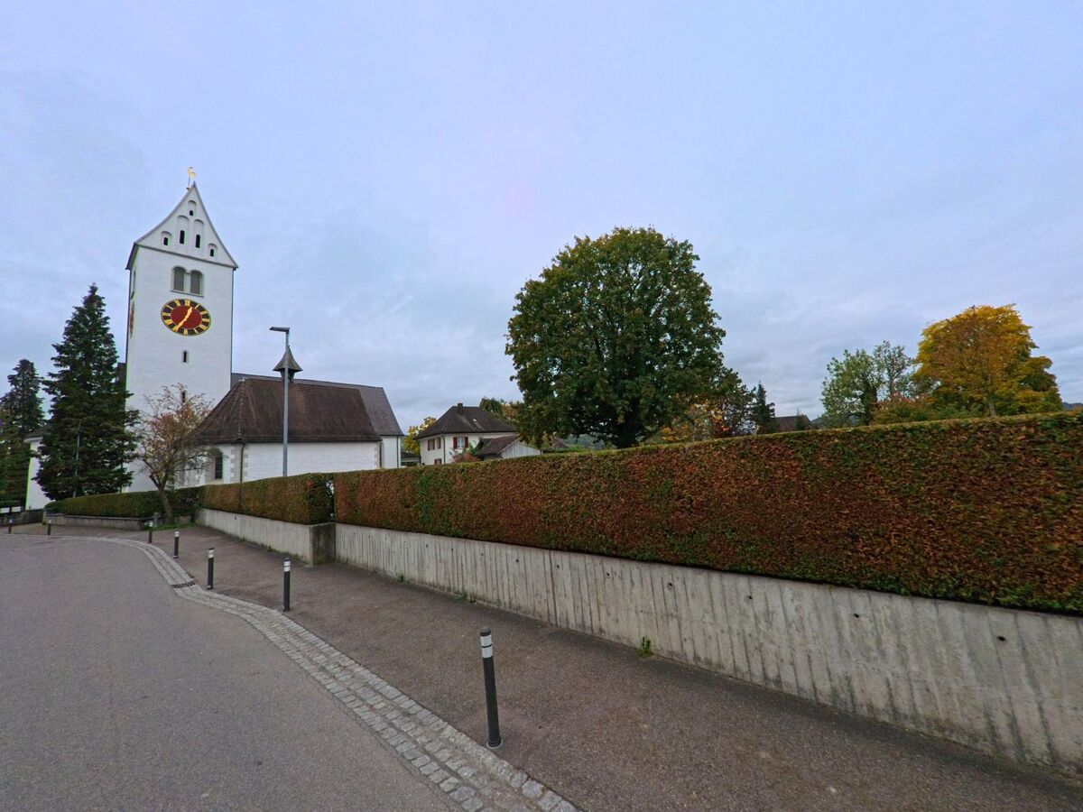 Entlang eines Gehwegs mit niedrigen Pollern erhebt sich der Kirchturm hinter einer Hecke im Ortskern von Wängi (9545, Kanton Thurgau) unter herbstlich truebem Himmel.