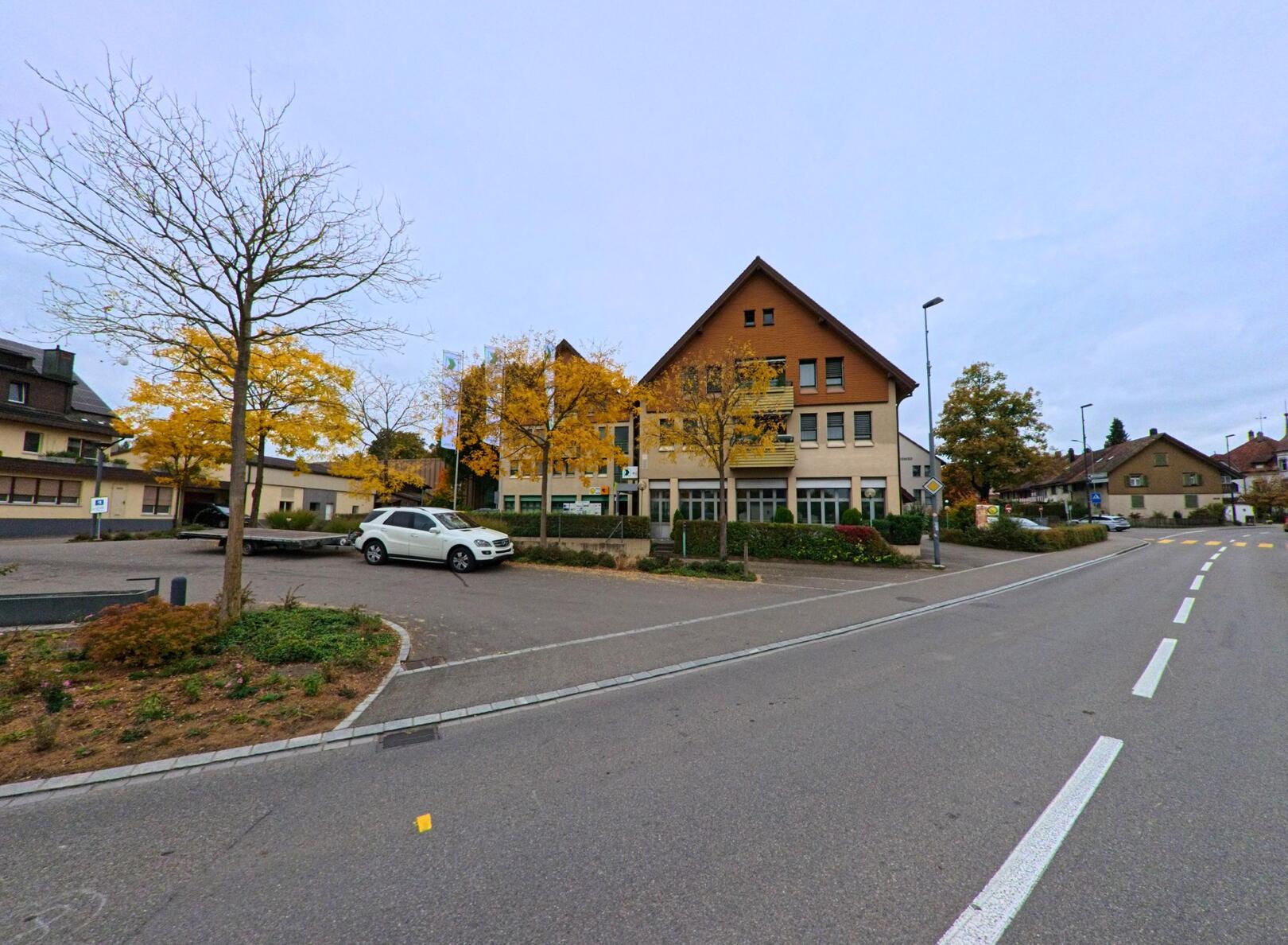 An einer ruhigen Strassenkreuzung in Wängi (9545, Kanton Thurgau) stehen Wohn- und Geschäftshäuser mit herbstlich gefärbten Bäumen unter klarem Himmel.