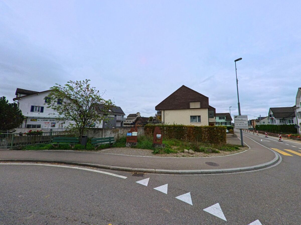Am Rand einer leicht gebogenen Strasse mit Sitzbank und Infotafeln zeigt sich ein ruhiger Wohnbereich in Wängi (9545, Kanton Thurgau) mit Einfamilienhaeusern und Gruenflaechen.