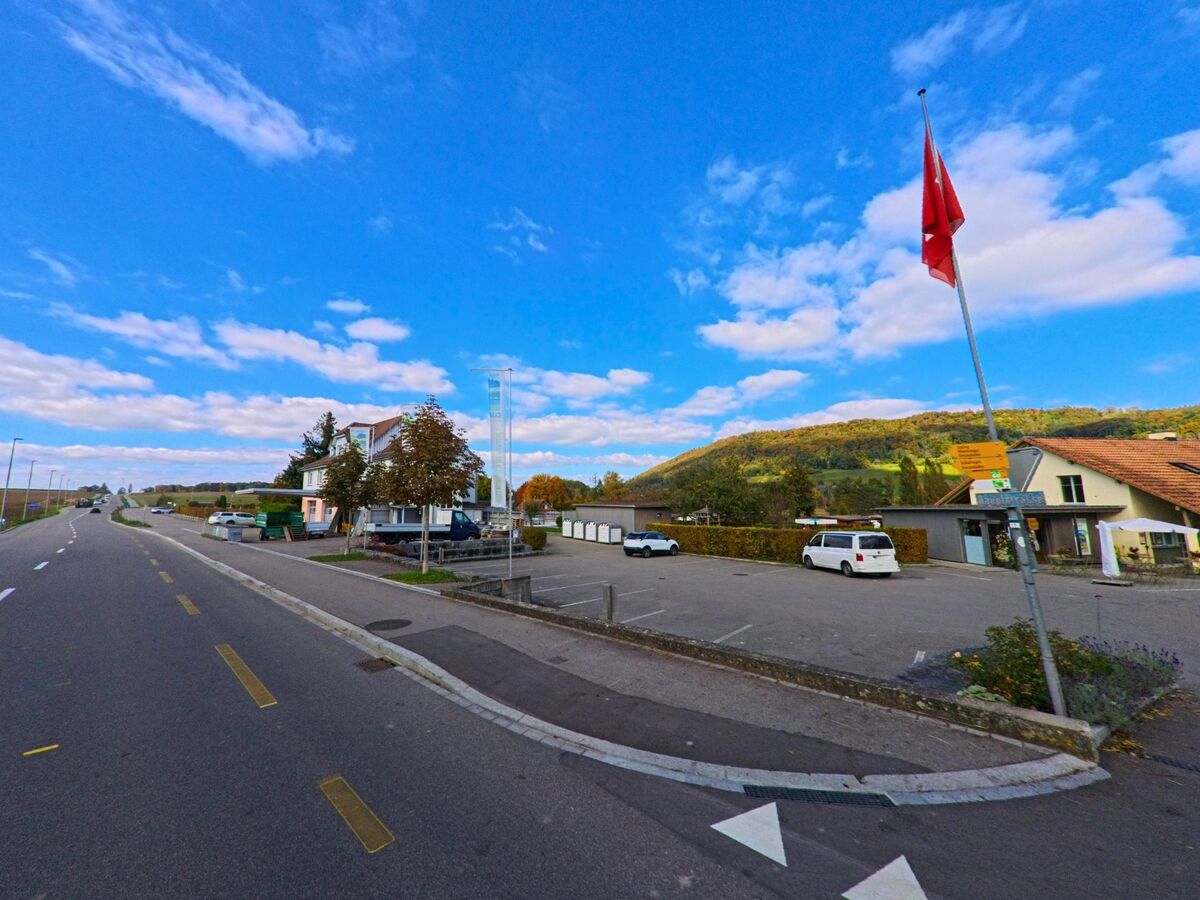 Am Rand einer breiten Strasse liegt in Wagenhausen (8259, Kanton Thurgau) ein offener Parkplatz mit Fahnenmast, umgeben von Gebaeuden und sanften Huegeln im Hintergrund.