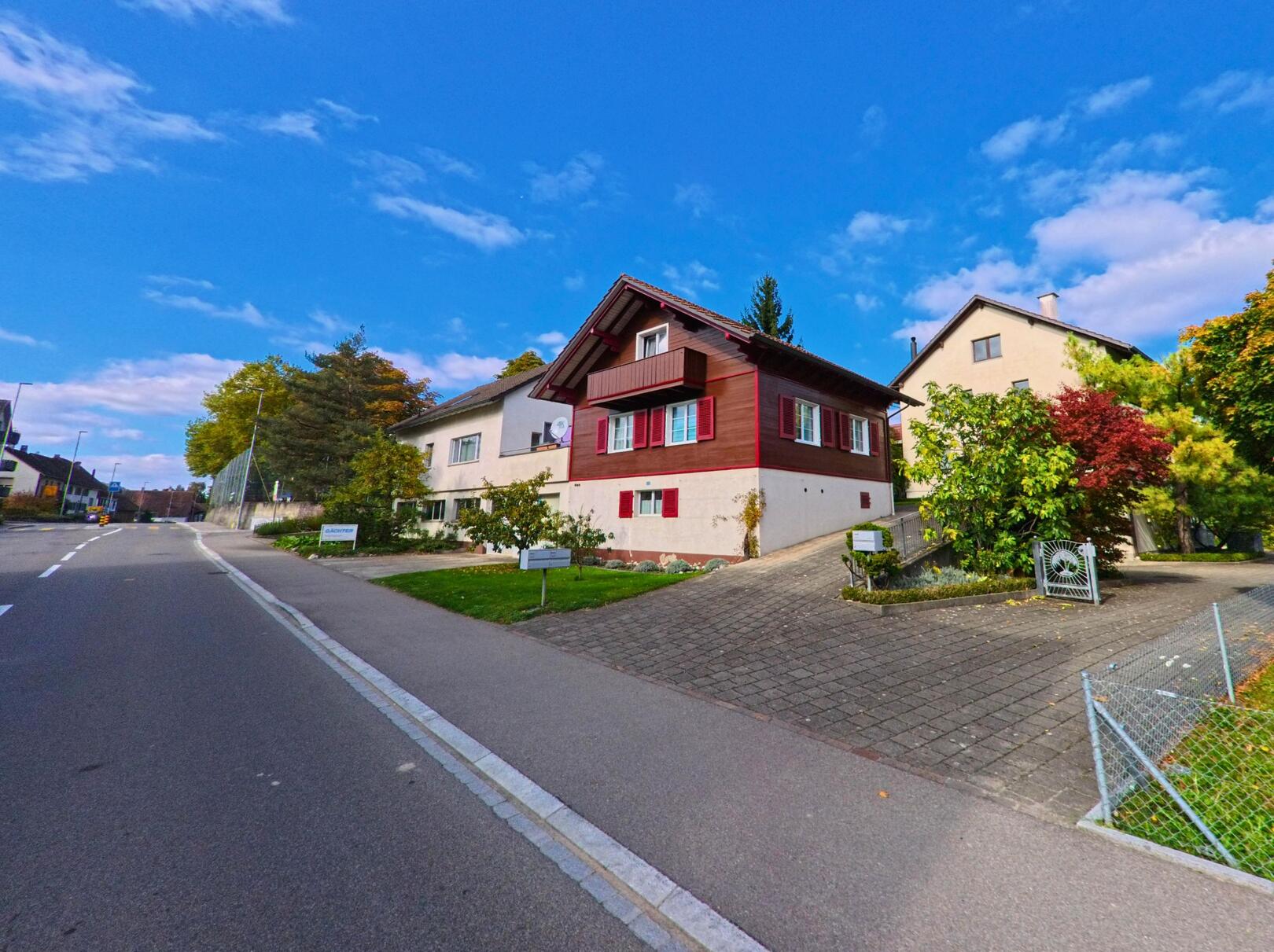 Entlang einer ruhigen Wohnstrasse in Wagenhausen (8259, Kanton Thurgau) stehen gepflegte Einfamilienhaeuser unter blauem Himmel mit herbstlicher Bepflanzung.