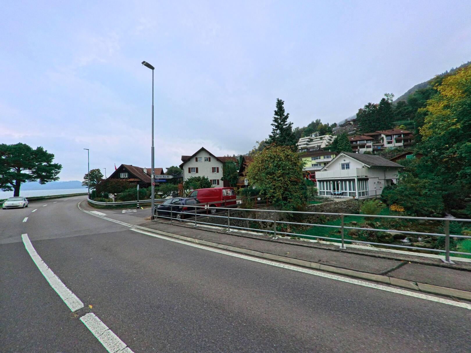 Ein modernes Wohnhaus in Walchwil (6318, Kanton Zug) steht an einer Strassenkurve, mit Blick auf den See und umgeben von Baeumen und anderen Wohngebaueten.