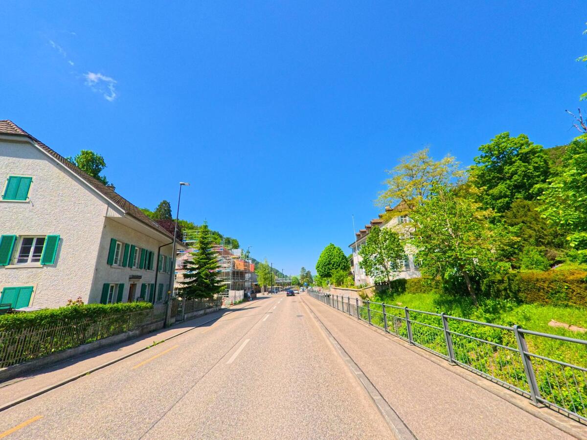  Eine breite Strasse durchquert Waldenburg (4437, Kanton Baselland), flankiert von Wohnhäusern mit grünen Fensterläden und einem Garten mit üppigem Baumbestand.