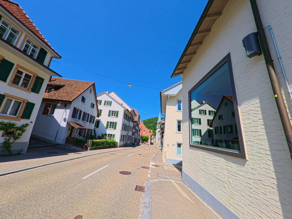 In Waldenburg (4437, Kanton Baselland) spiegeln sich historische Fassaden mit grünen Fensterläden in einem modernen Schaufenster entlang einer ruhigen Strasse.