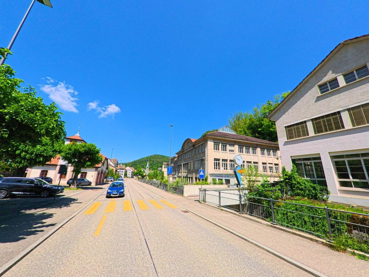 In Waldenburg (4437, Kanton Baselland) verläuft eine breite Strasse mit Fussgängerstreifen entlang historischer Industriegebäude und eines kleinen Bahnhofs mit Uhr.