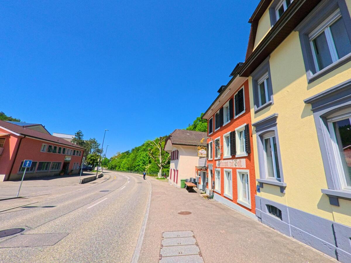  In Waldenburg (4437, Kanton Baselland) verläuft eine Strasse vorbei an farbig gestrichenen Gebäuden, darunter das traditionsreiche Gasthaus „Zur Post“, mit Blick ins Grüne.