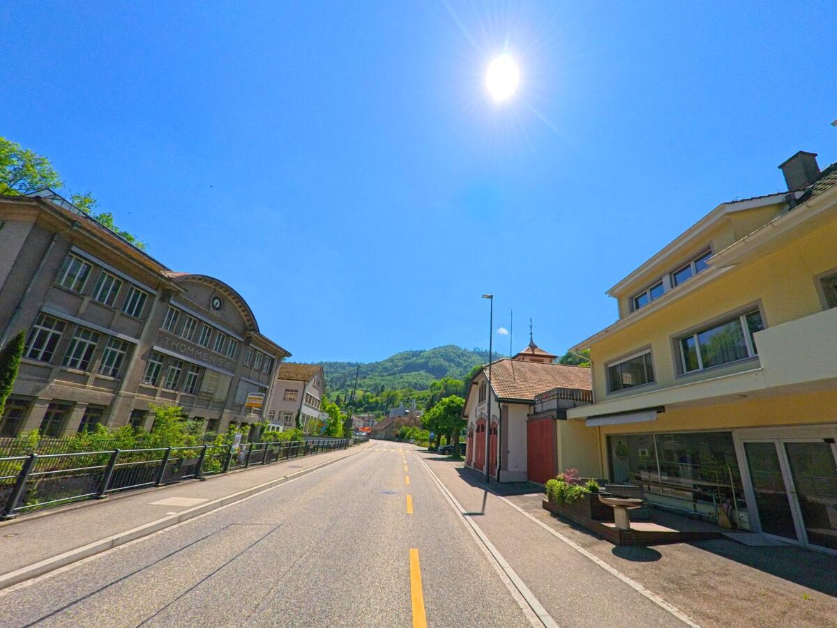  In Waldenburg (4437, Kanton Baselland) verläuft eine breite Strasse vorbei an einem historischen Fabrikgebäude mit Uhr und modernen Wohnhäusern unter blauem Himmel.