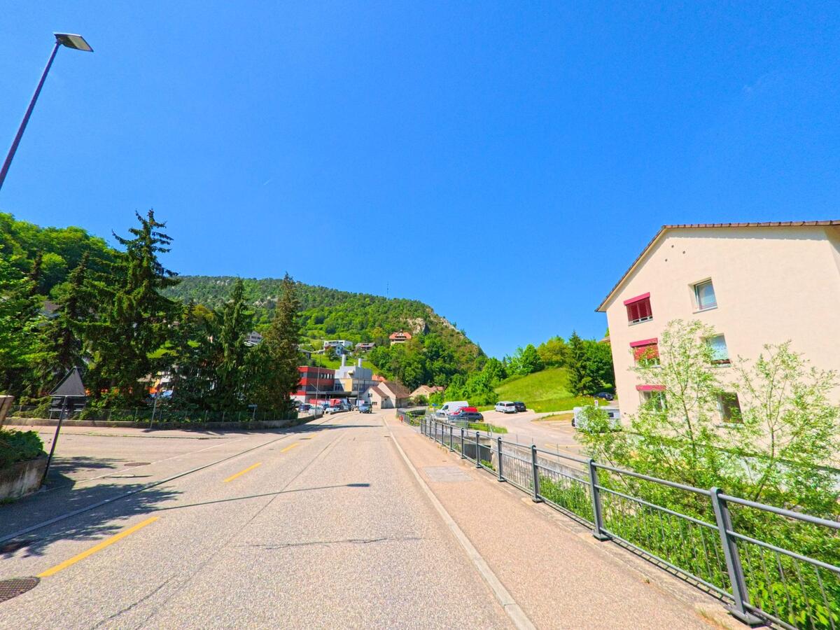 Am Ortsrand von Waldenburg (4437, Kanton Baselland) führt eine Strasse an einem Wohnhaus mit roten Fensterläden vorbei in Richtung bewaldeter Hanglage mit verstreuten Häusern.