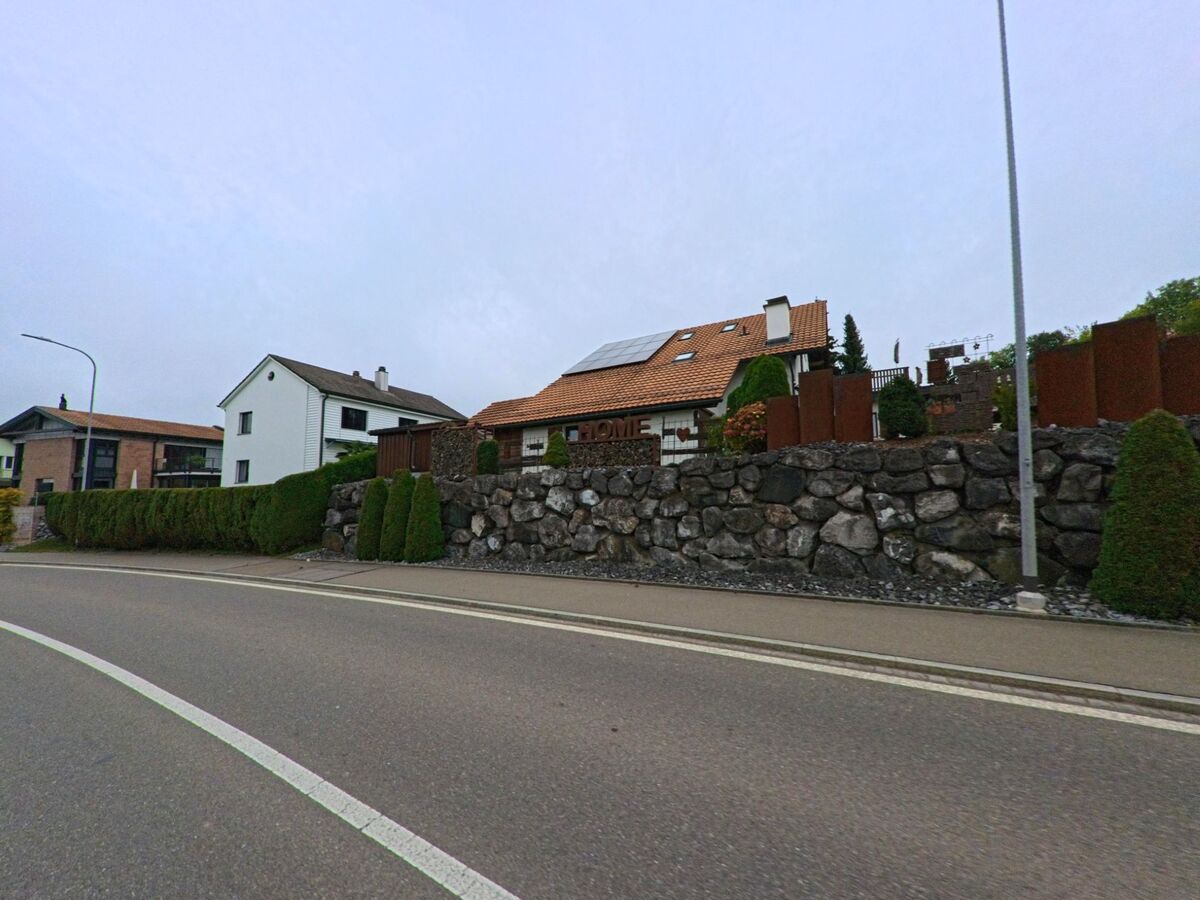 Entlang einer Ortsstrasse in Waldkirch (9205, Kanton St. Gallen) liegen Einfamilienhaeuser hinter einer Natursteinmauer mit gepflegten Gaerten.