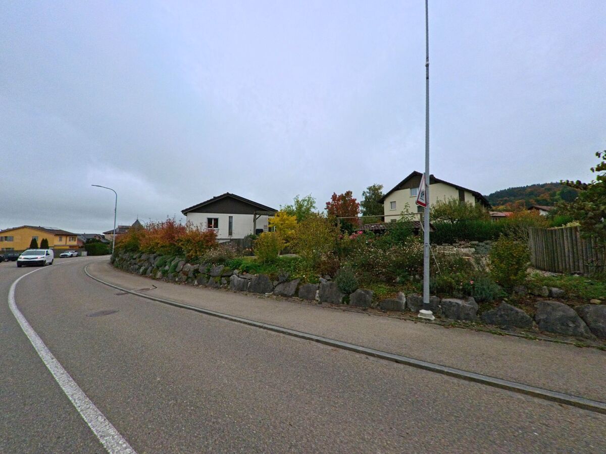 Entlang einer leicht geschwungenen Strasse in Waldkirch (9205, Kanton St. Gallen) verlaufen Einfamilienhaeuser hinter einer bepflanzten Steinboeschung.