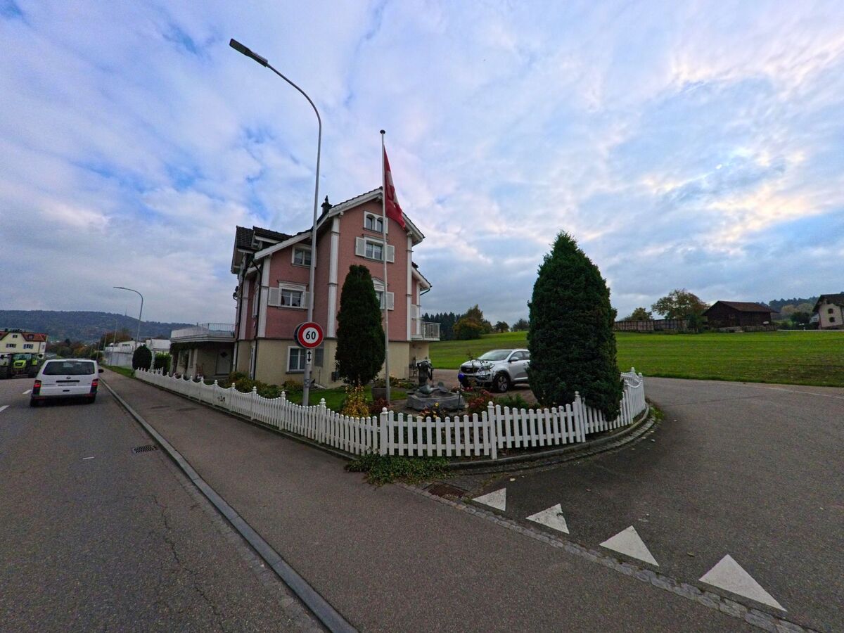 An einer Strassenkreuzung in Waldkirch (9205, Kanton St. Gallen) steht ein Wohnhaus mit Schweizer Fahne, umgeben von Zaun, Garten und offener Gruenflaeche.