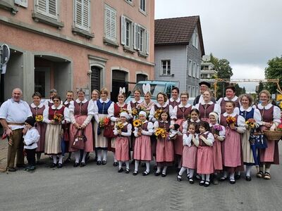 In Einsiedeln (8842, Kanton Schwyz) steht die Gruppe Waldluet vo Einsidle in traditionellen Trachten mit Blumenstraeussen vor Wohnhaeusern zusammen.
