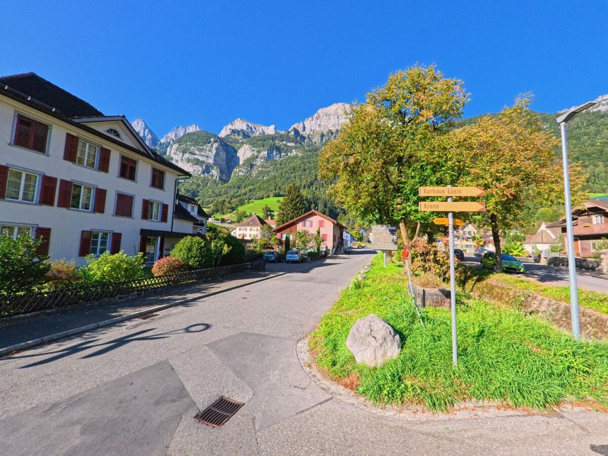 Eine ruhige Strasse in Walenstadt (8880, Kanton St. Gallen) führt vorbei an gepflegten Häusern und Wegweisern zum Kurhaus Lusis, mit den steilen Felsen der Churfirsten im Hintergrund.