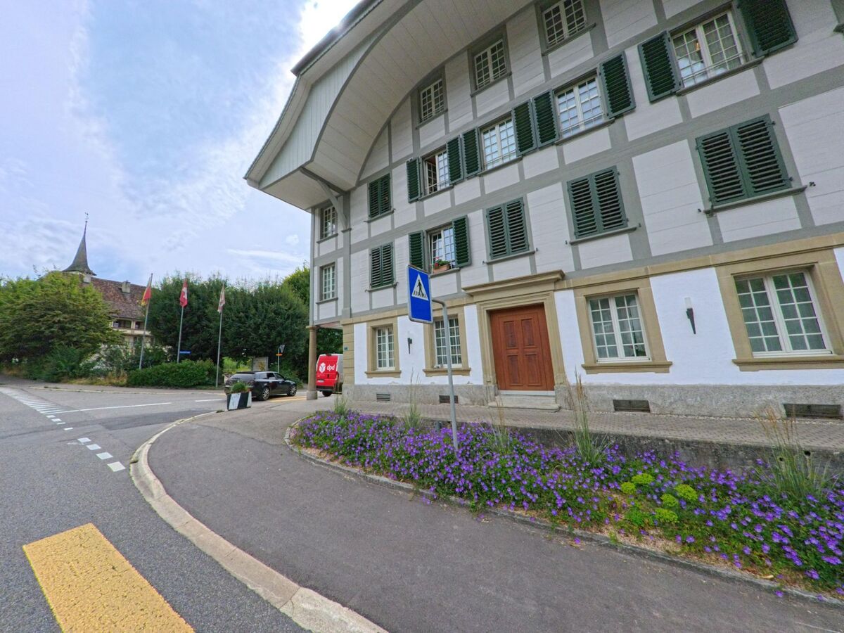 Vor einem stattlichen Gebäude mit grün-weissen Fensterläden in Walkringen (3512, Kanton Bern) blühen violette Blumen, im Hintergrund ragt der Kirchturm empor.