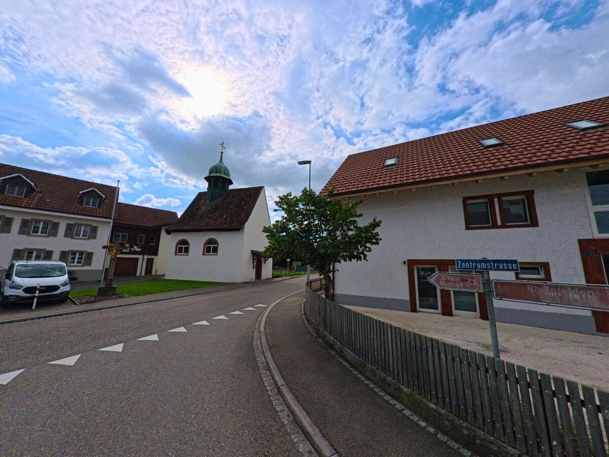 Eine kleine Kapelle mit Zwiebelturm steht an der Zentrumstrasse in Wallbach (4323, Kanton Aargau), eingerahmt von Wohnhaeusern und leicht bewölktem Himmel.