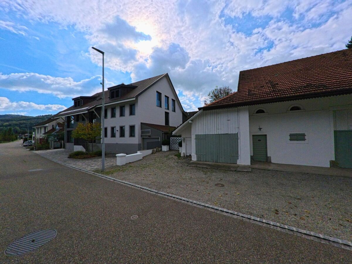 Entlang einer ruhigen Strasse in Wallbach (4323, Kanton Aargau) stehen moderne Wohnhaeuser neben einem traditionellen Gebaeude mit gruener Tuere.