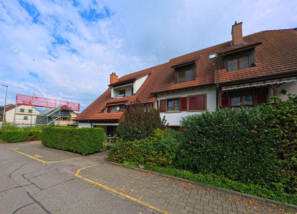 Vor einem grossen Wohnhaus mit roten Fensterlaeden in Wallbach (4323, Kanton Aargau) stehen dichte Hecken, waehrend links Baugerueste sichtbar sind.