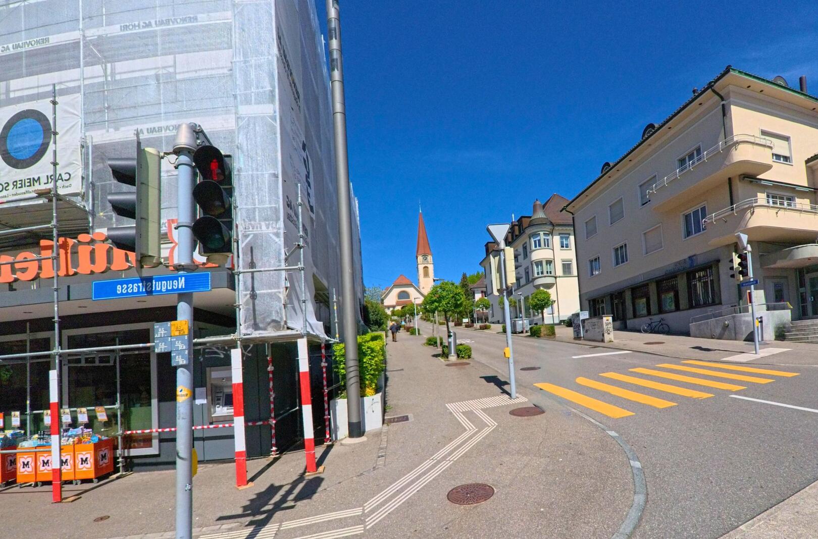 In Wallisellen (8304, Kanton Zürich) überblickt man von der Neugutstrasse eine Kreuzung mit Geschäften, Fussgängerstreifen und einer Kirche im Hintergrund.