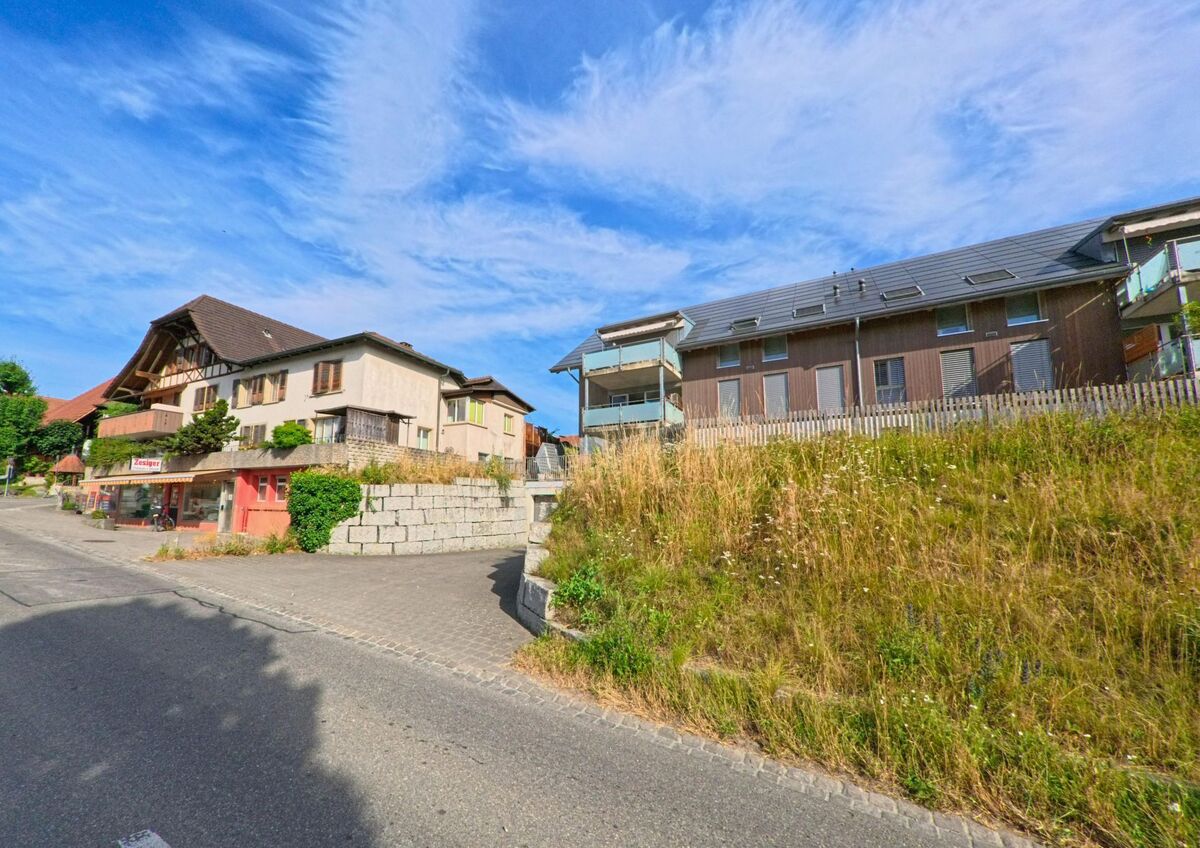 In Walperswil (3275, Kanton Bern) stehen entlang einer leicht ansteigenden Strasse ein altes Wohnhaus mit Ladengeschäft und ein modernes Mehrfamilienhaus mit Balkonen.