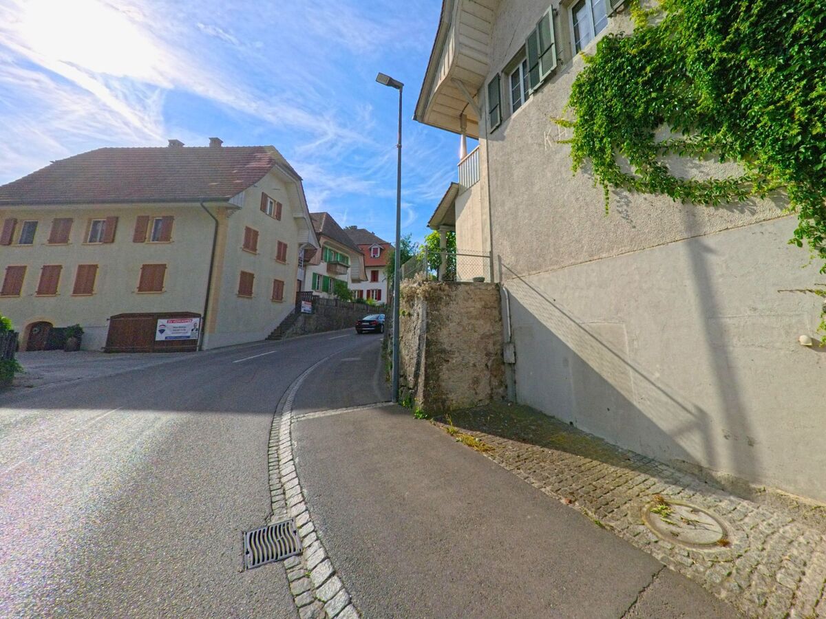 In Walperswil (3275, Kanton Bern) verläuft eine enge Strasse mit leichten Steigungen zwischen historischen Wohnhäusern mit Fensterläden und begrünter Fassade.