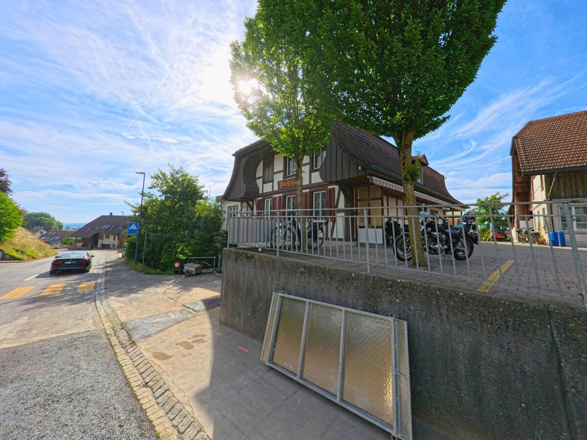 In Walperswil (3275, Kanton Bern) liegt ein traditionelles Gebäude mit Fachwerkfassade an einer Hangstrasse, davor parken Motorräder im Schatten eines Baumes.