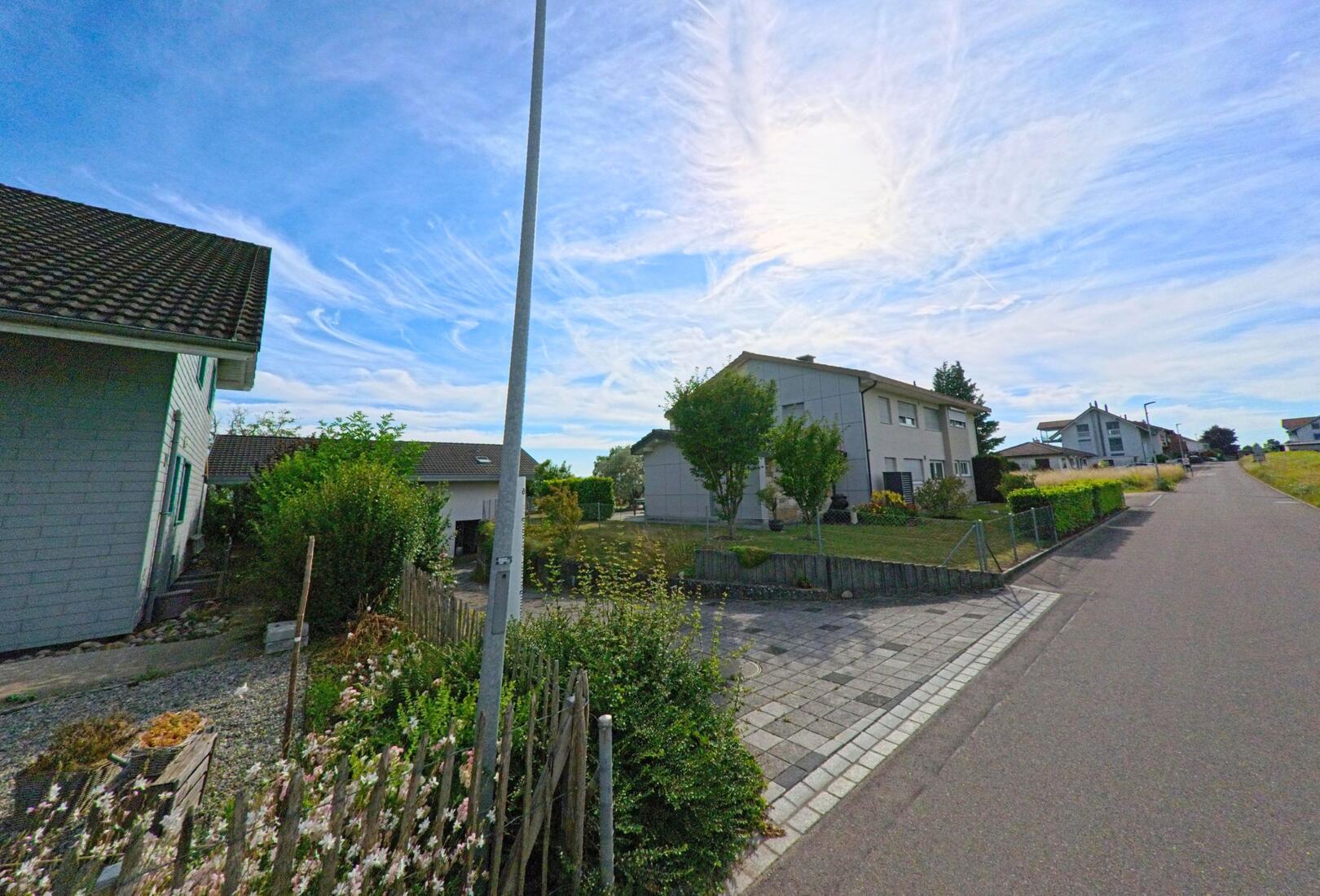 In Walperswil (3275, Kanton Bern) verläuft eine ruhige Quartierstrasse mit Einfamilienhäusern, kleinen Vorgärten und einem einzelnen Laternenmast unter sommerlichem Himmel.
