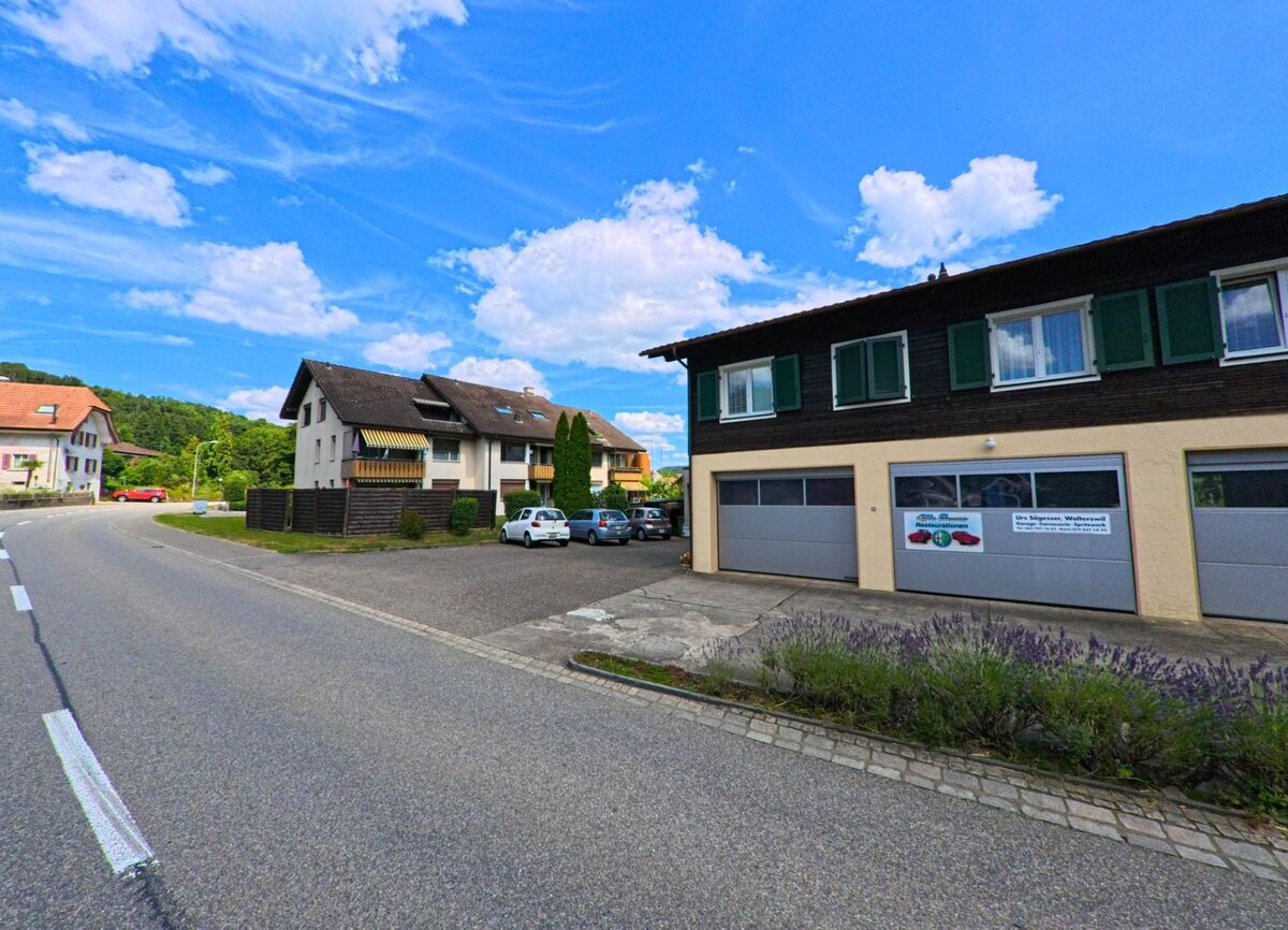 An einer leicht geschwungenen Strasse in Walterswil (5746, Kanton Solothurn) befindet sich eine kleine Autowerkstatt im Erdgeschoss eines Hauses mit grünen Fensterläden, gegenüber mehrstöckigen Wohnhäusern.