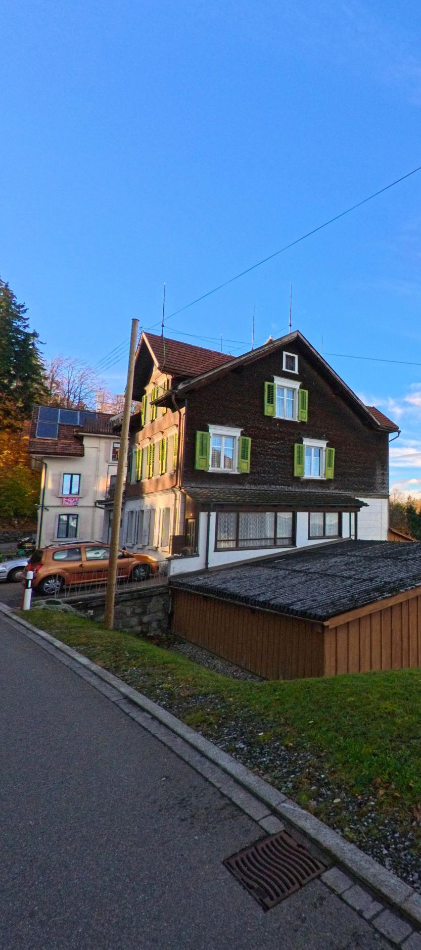 Ein traditionelles Holzhaus mit gruene Fensterlaeden steht in Walzenhausen (9428, Kanton Appenzell Ausserrhoden) am Strassenrand unter klarem Abendhimmel.