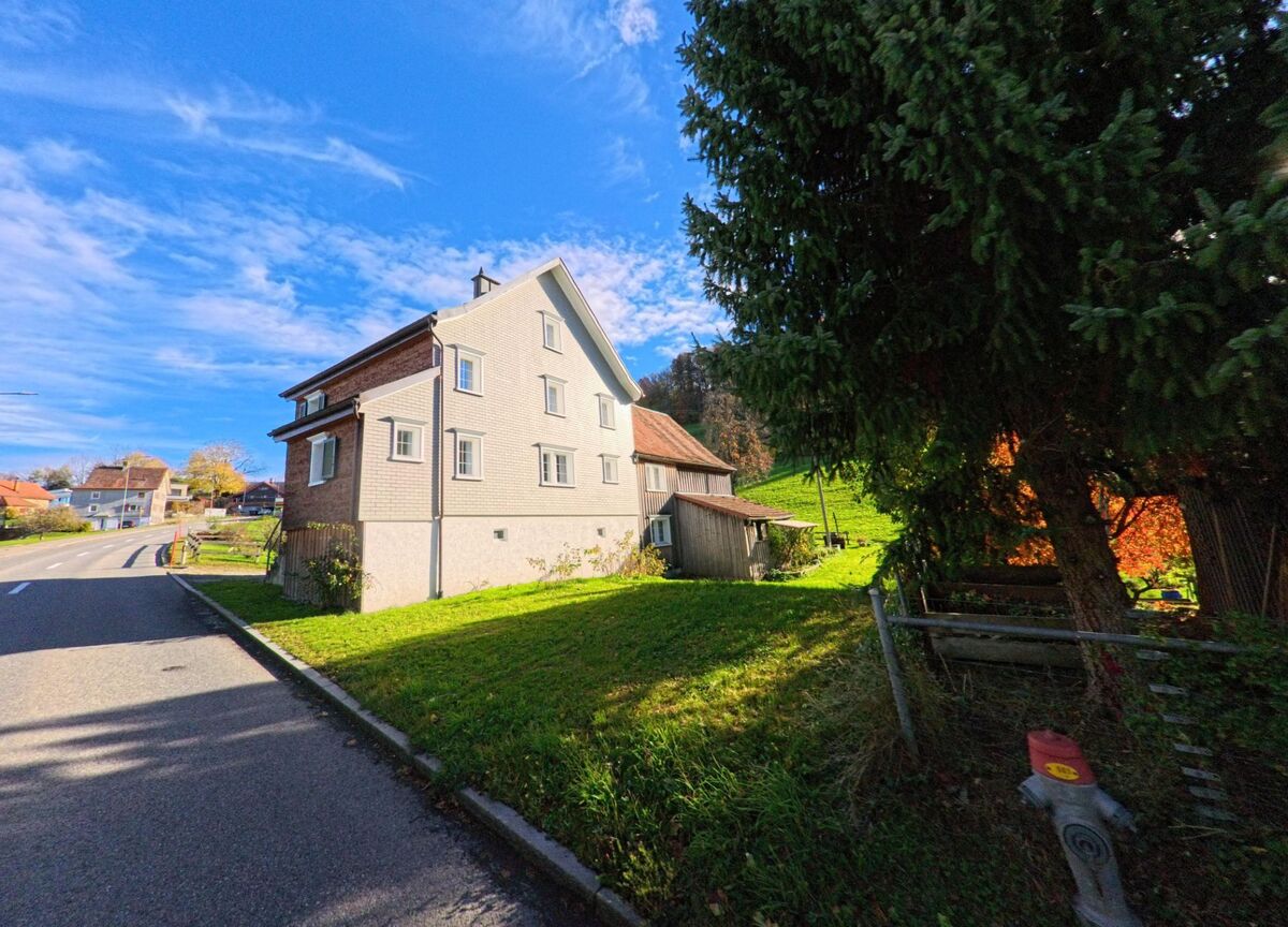 Ein freistehendes Wohnhaus steht in Walzenhausen (9428, Kanton Appenzell Ausserrhoden) am sonnigen Hang neben einer Strasse, waehrend dahinter gruene Wiesen ansteigen.