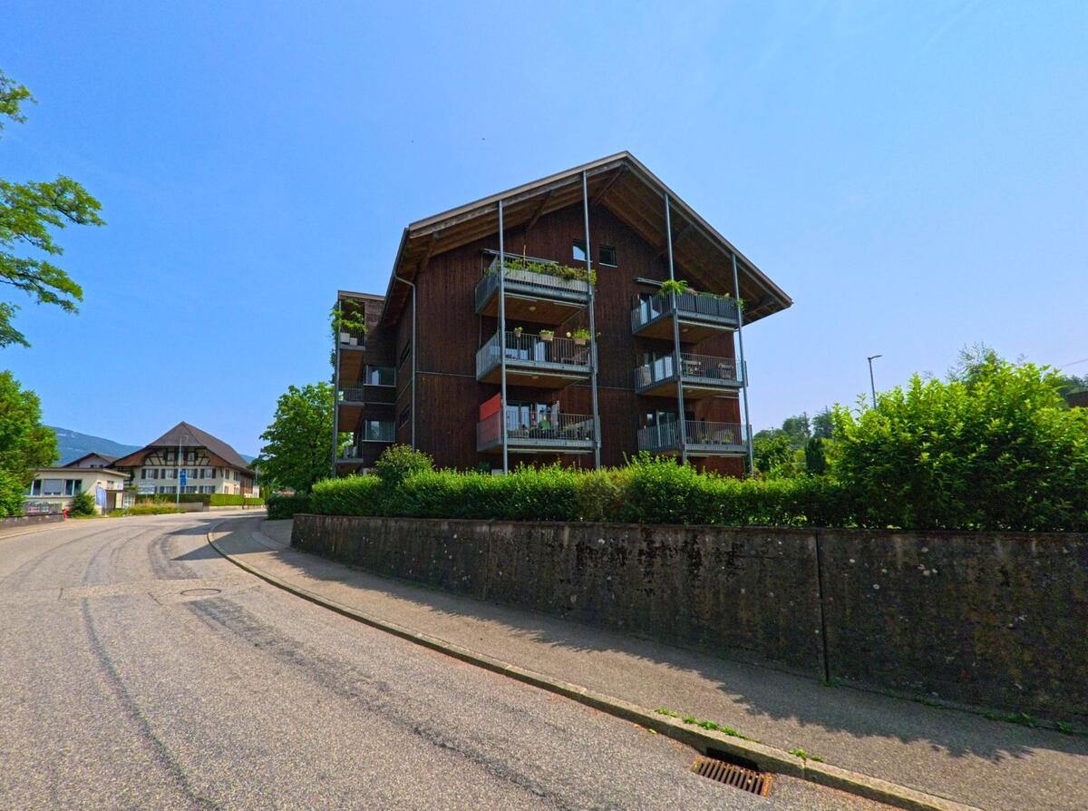 Ein modernes Mehrfamilienhaus in Holzbauweise steht in Wangen an der Aare (3380, Kanton Bern) an einer Kurve mit Blick auf traditionelle Gebäude im Hintergrund.