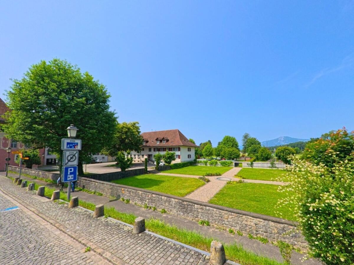 In Wangen an der Aare (3380, Kanton Bern) liegt ein gepflegter Grünbereich mit Wegen und Ziersträuchern vor historischen Gebäuden und mit Blick auf den Jura.