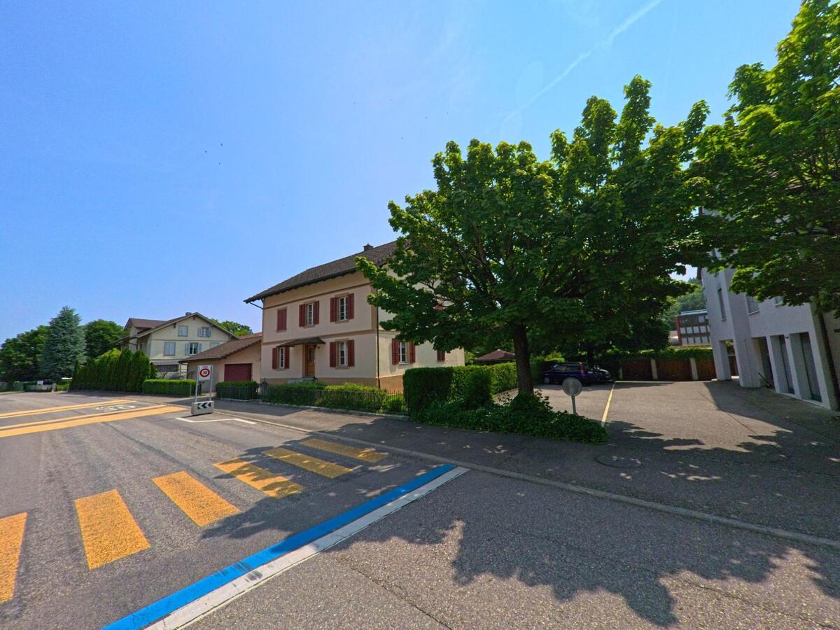 An einer verkehrsberuhigten Strasse in Wangen an der Aare (3380, Kanton Bern) steht ein Wohnhaus mit roten Fensterläden neben einem schattigen Parkplatz.
