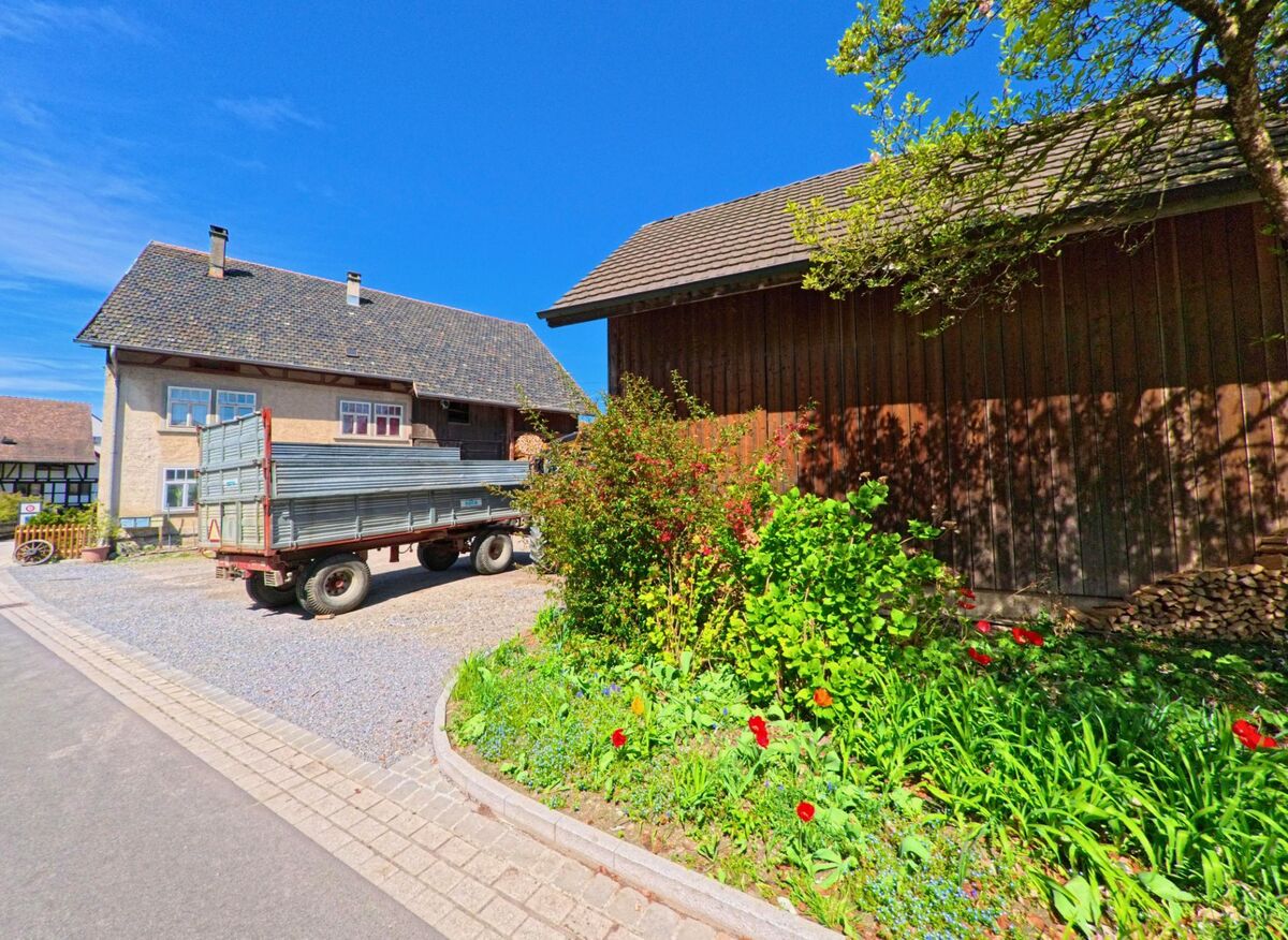 In Wangen-Brüttisellen (PLZ 8602, Kanton Zürich) steht ein Anhänger auf einem Hofplatz zwischen einem Wohnhaus und einer Holzscheune, umgeben von Blumen und Sträuchern.