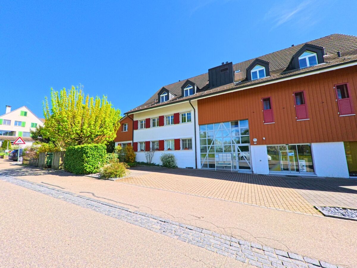 In Wangen-Brüttisellen (8602, Kanton Zürich) steht ein modernes Gebäude mit grossflächiger Fensterfront und roten Fensterläden an einer ruhigen Strasse mit Baumallee.