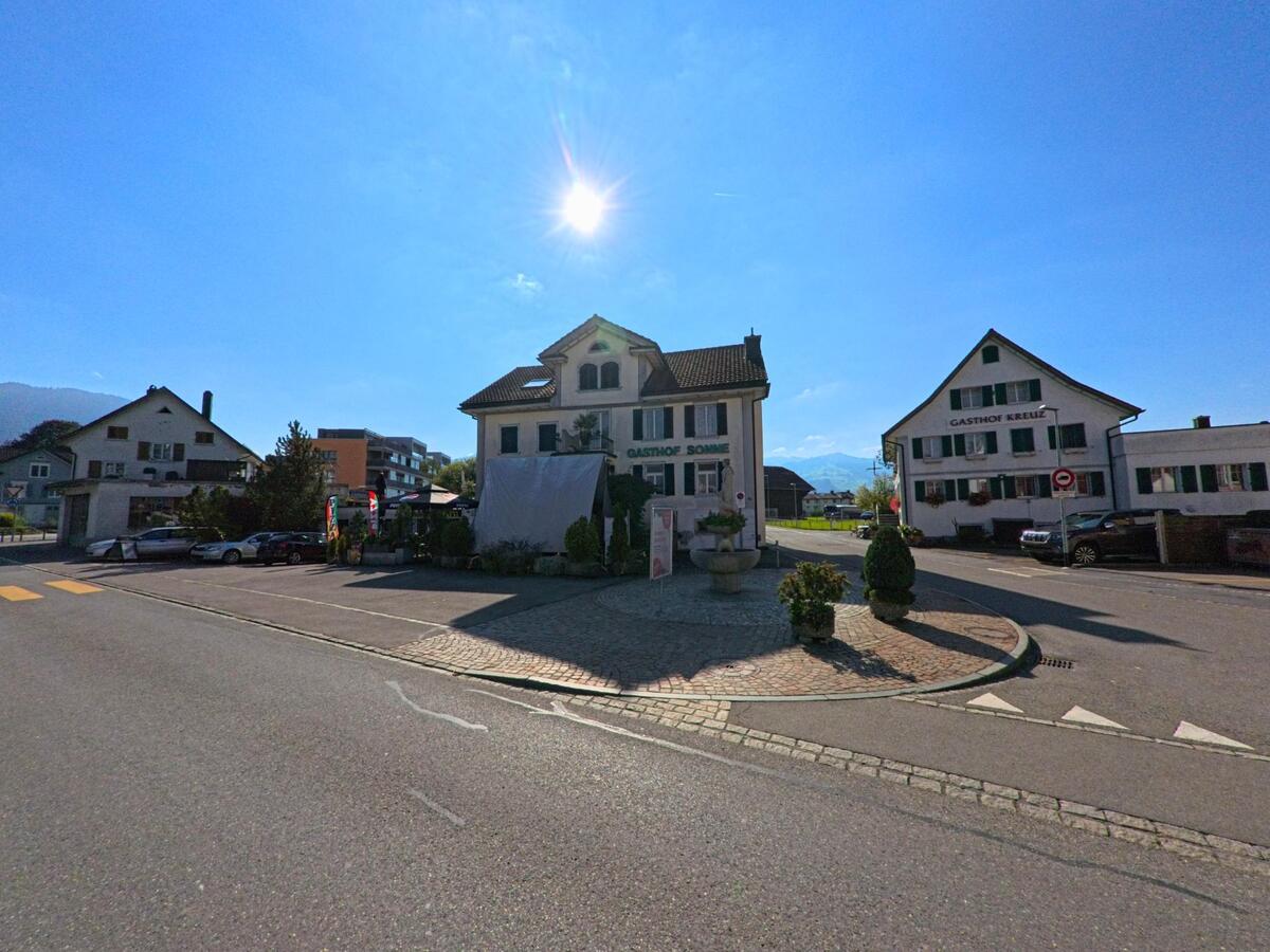 Zwei traditionelle Gasthoefe in Wangen_(SZ) (8855, Kanton Schwyz) stehen sich an einer sonnigen Strassenkreuzung gegenueber, waehrend im Hintergrund Berge erkennbar sind.