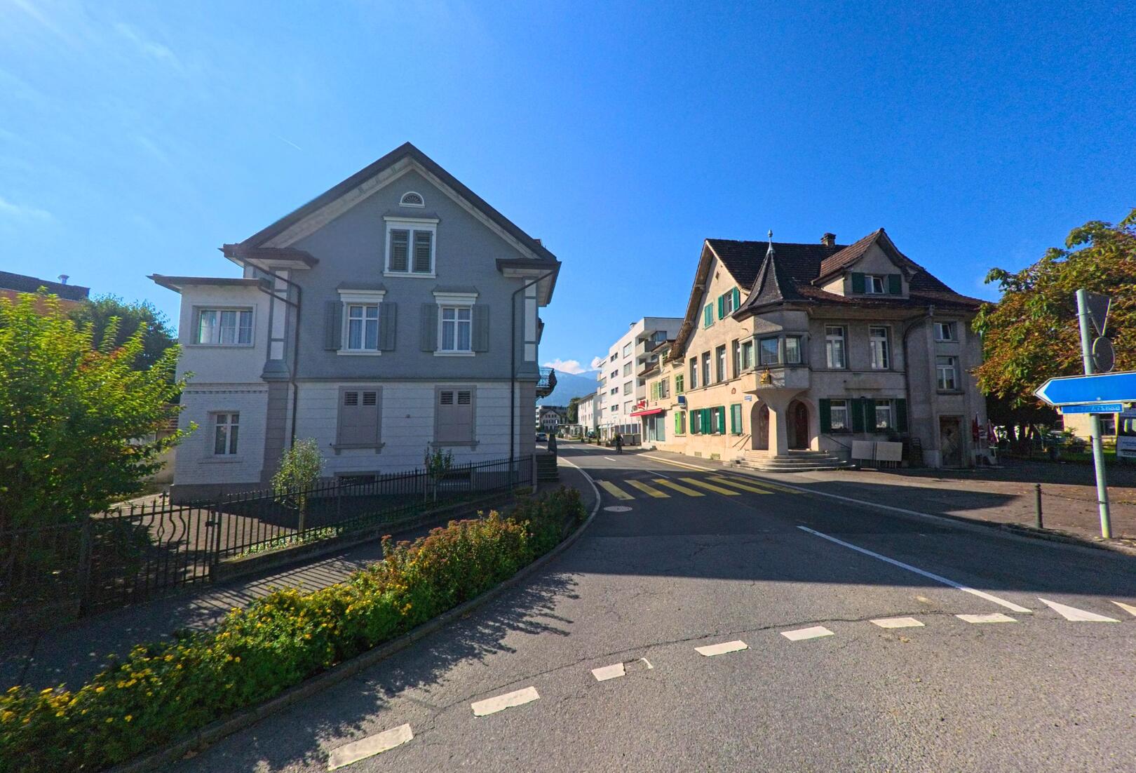 In Wangen_(SZ) (8855, Kanton Schwyz) zeigt das Bild eine sonnige Strassenszene mit zwei traditionellen Wohnhaeusern, die sich an einer leichten Kurve gegenueberstehen.