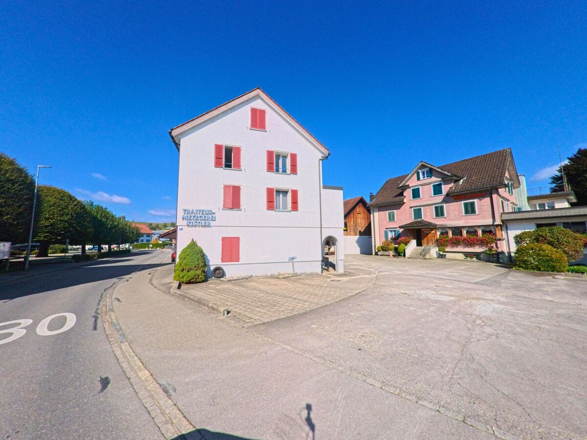 In Wangen_(SZ) (8855, Kanton Schwyz) stehen zwei Wohnhaeuser mit farbigen Fensterlaeden an einer leicht geschwungenen Strasse, die von Baeumen gesaeumt wird.