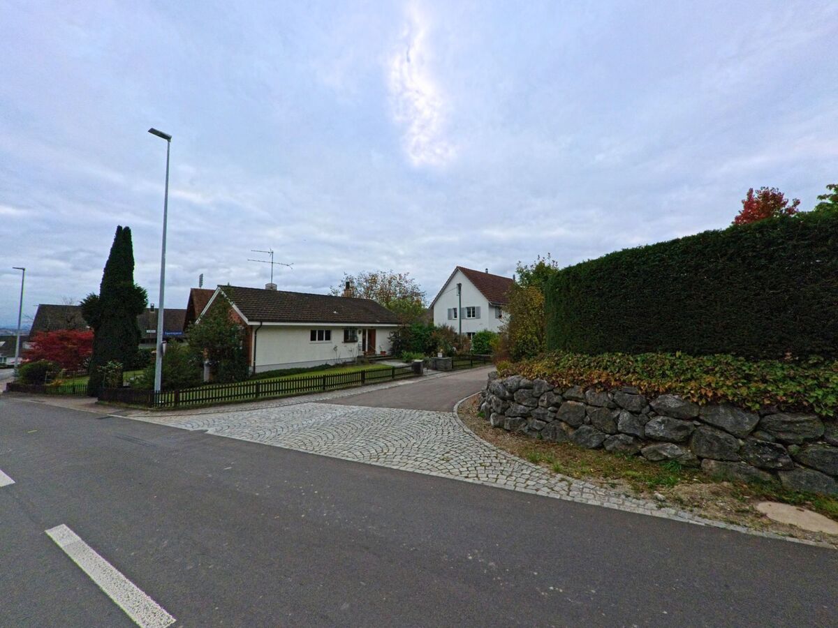 An einer ruhigen Einmündung mit Natursteinmauer und gepflegten Vorgärten liegen Einfamilienhäuser in Warth-Weiningen (8532, Kanton Thurgau) unter leicht bewölktem Himmel.