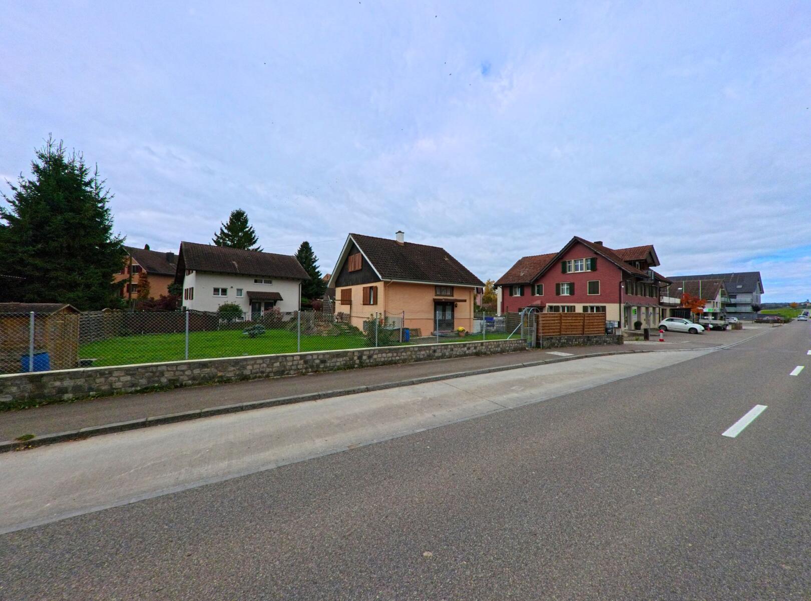 Entlang einer ruhigen Wohnstrasse stehen Einfamilienhaeuser mit Gaerten in Warth-Weiningen (8532, Kanton Thurgau) unter einem leicht bewoelkten Himmel.